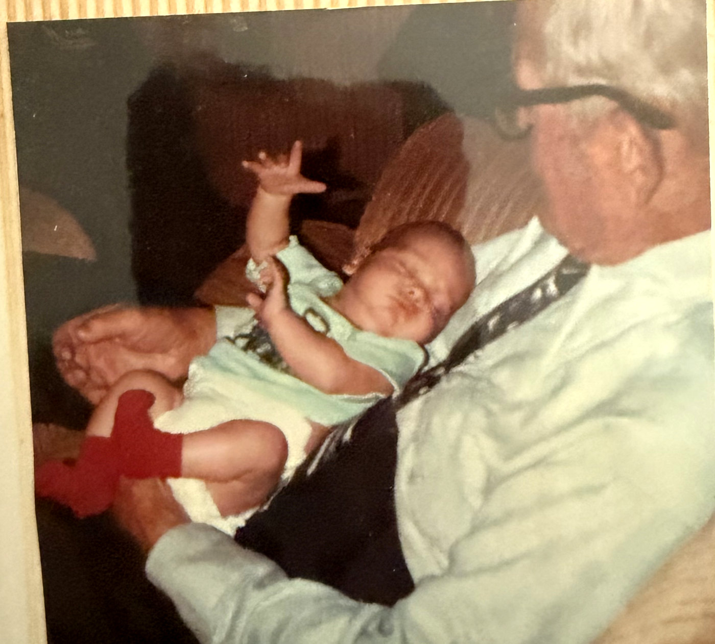 A faded colour photograph of an elderly William Thompson Hanes, in glasses, shirt and tie, holding his newborn great-grandson in his arms.
