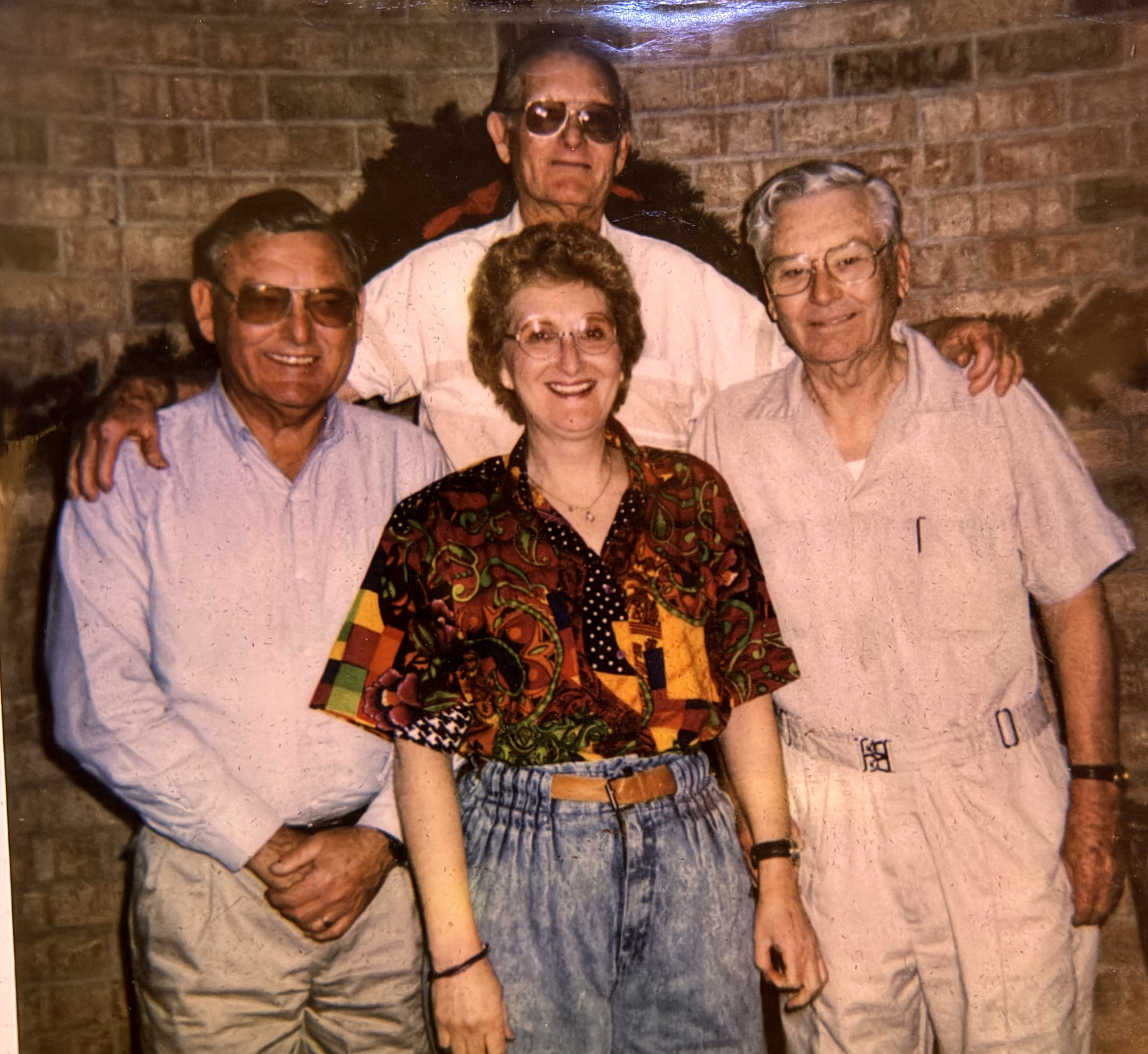 A colour photograph of the four Hanes siblings together in later life, standing in front of a brick wall. Three men and one woman, all wearing glasses.